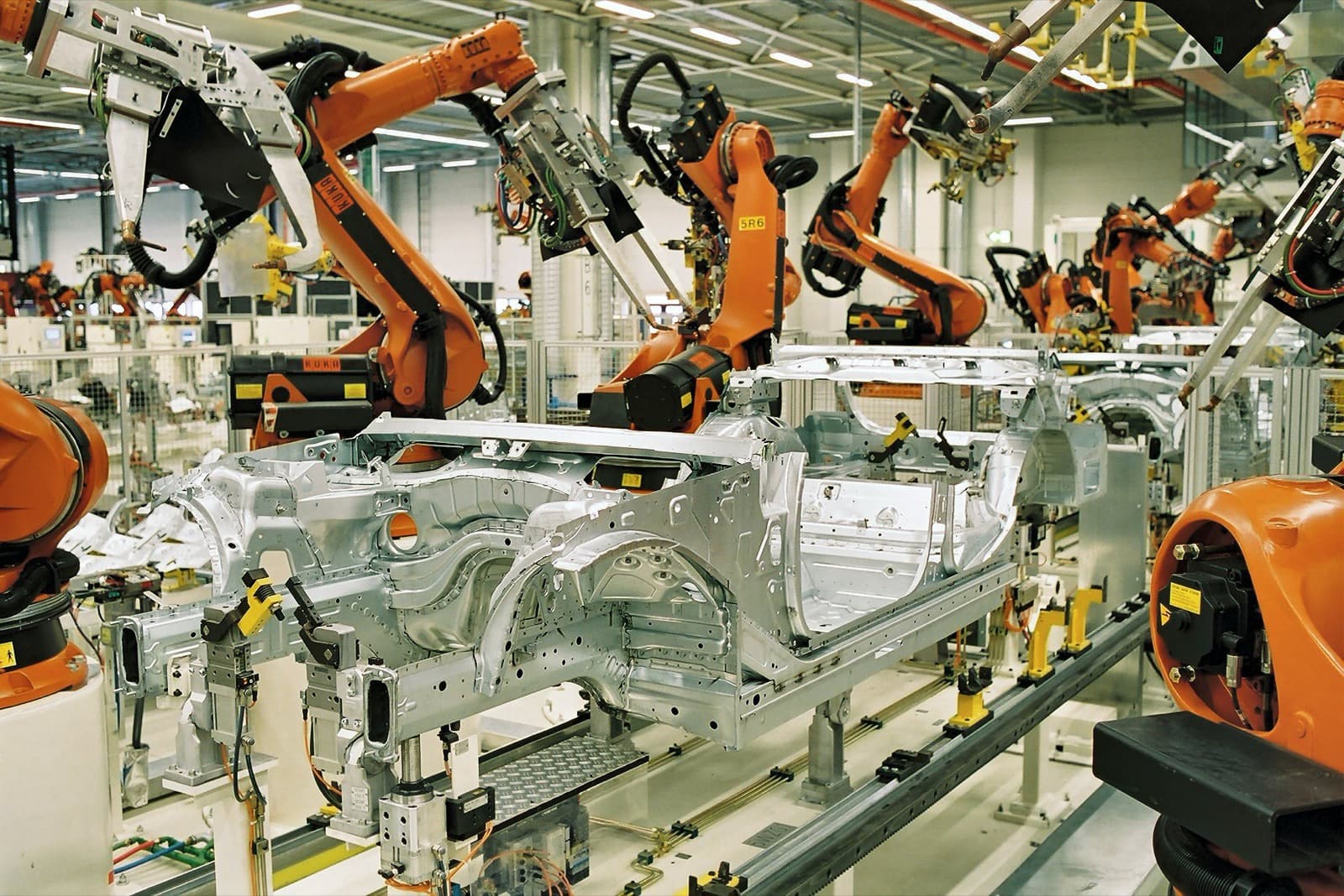 Orange industrial robot arms welding silver car bodies on an automotive assembly line.